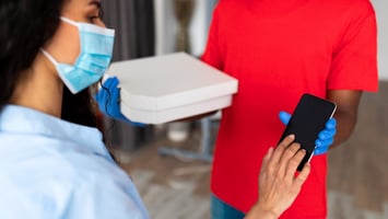 Persona con mascarilla recibe un pedido de un repartidor con guantes y uniforme rojo, confirmando la entrega en un teléfono móvil.