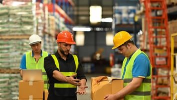 Tres trabajadores de almacén con cascos de seguridad y chalecos reflectantes revisan cajas. Dos de ellos inspeccionan un paquete en primer plano, mientras que otro utiliza una laptop al fondo, rodeado de pallets apilados.