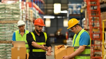 Trabajadores con casco y chaleco de seguridad revisando paquetes en un almacén logístico, uno de ellos usando computadora portátil.