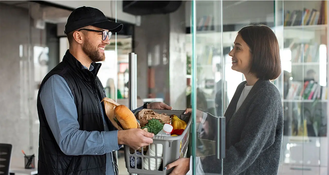 Repartidor con gorra y chaleco entregando una cesta con verduras, pan y lácteos a una mujer a través de una puerta de cristal.