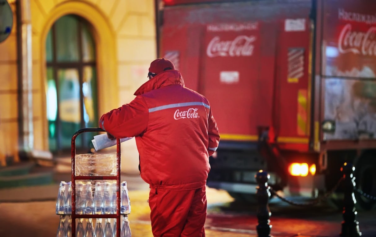 Repartidor de Coca-Cola con uniforme rojo descarga botellas de vidrio desde camión de reparto en zona urbana.