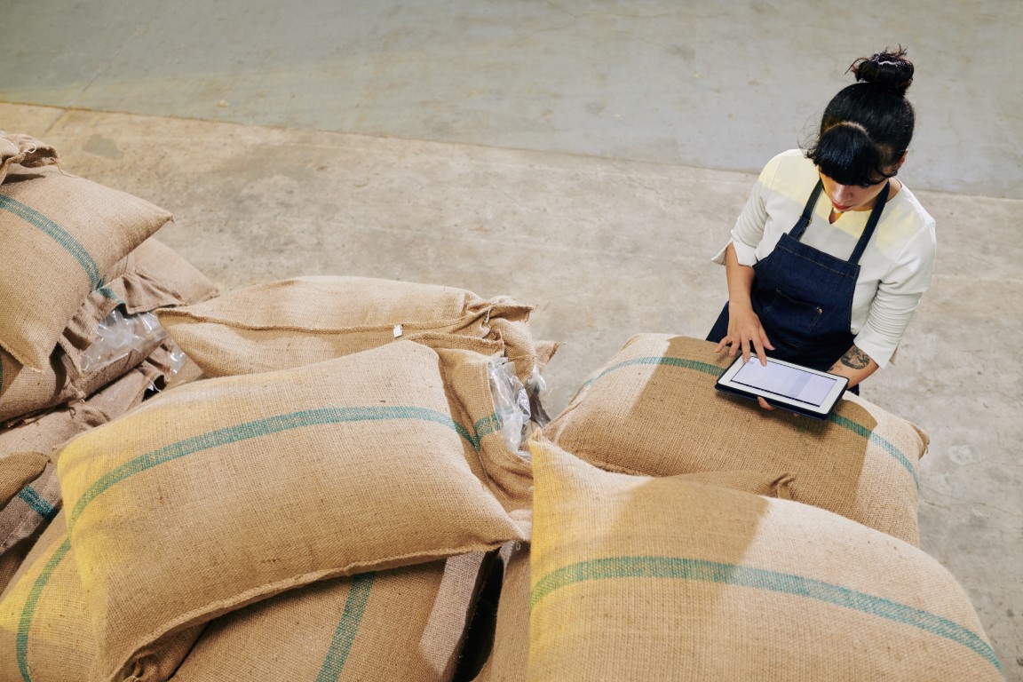 Trabajadora con tableta digital revisando inventario de sacos de granos en almacén logístico.